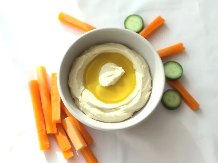 Cuenco de hummus con aceite por encima, rodeado de bastones de zanahoria y pepino